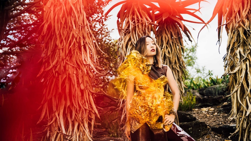 In this red-hued photo, an Asian woman in a modern yellow dress is standing defiantly in front of tall tress