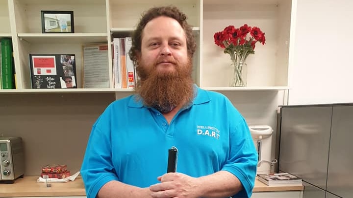 A man with ginger hair and a long beard stands in an office, holding a cane.