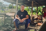 Young man sits in an open shed wearing Samoan ie lavalava, smiles as he looks to his right. 