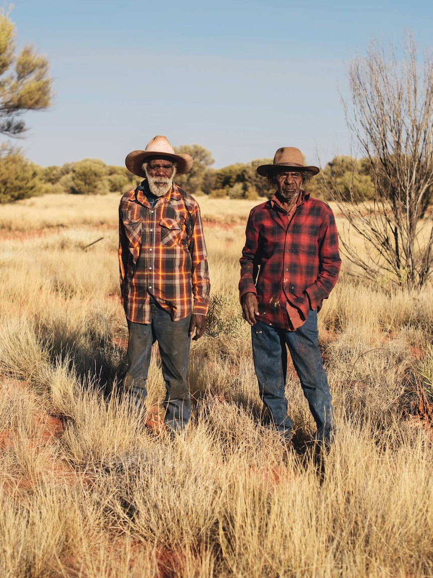 Two aboriginal men stand in flannelette shirts among dried yellow grass, red dirt and green trees