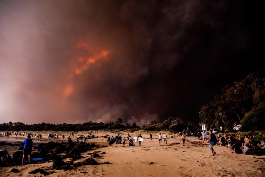 People on beach with large fire visible through dark smoke in background