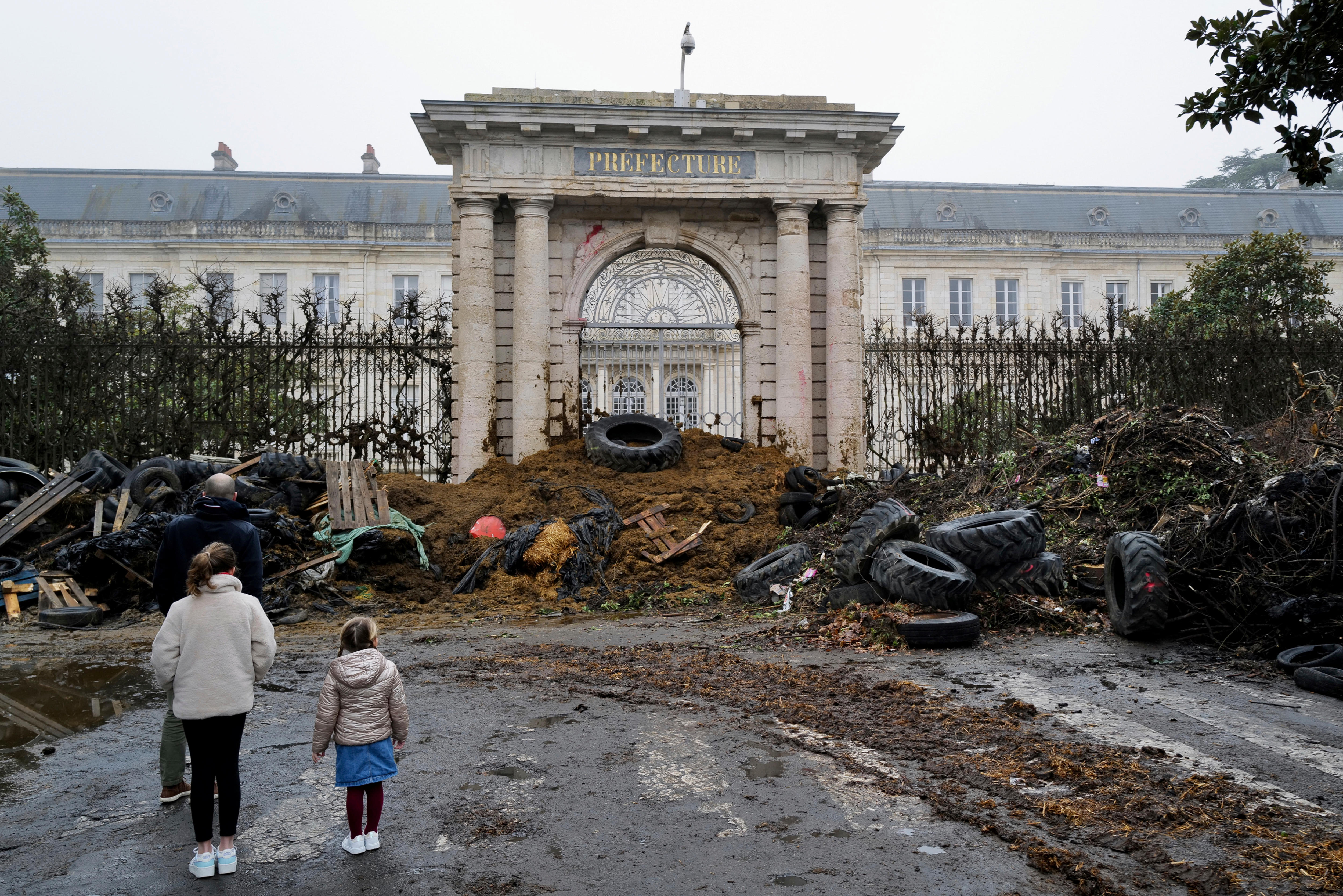 Two people stand in front of manure and tyres stacked in a heap at the gates of a large building.
