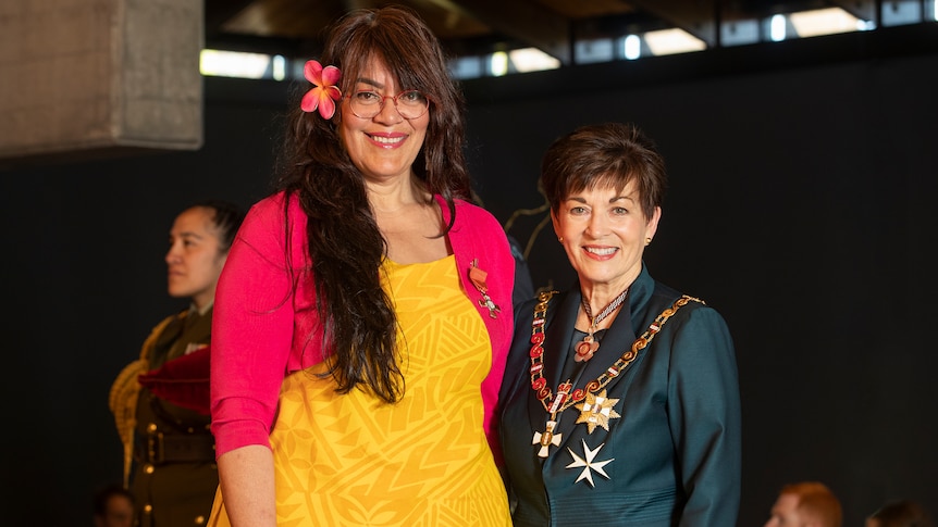 Pacific woman with long hair and flower in ear wears yellow dress and stands next to another shorter woman in navy shirt medals.