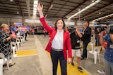 A woman in a red jacket waves to camera and fans in a warehouse.
