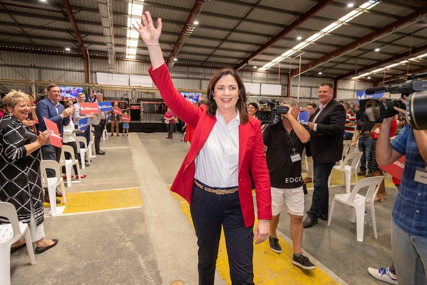 A woman in a red jacket waves to camera and fans in a warehouse.