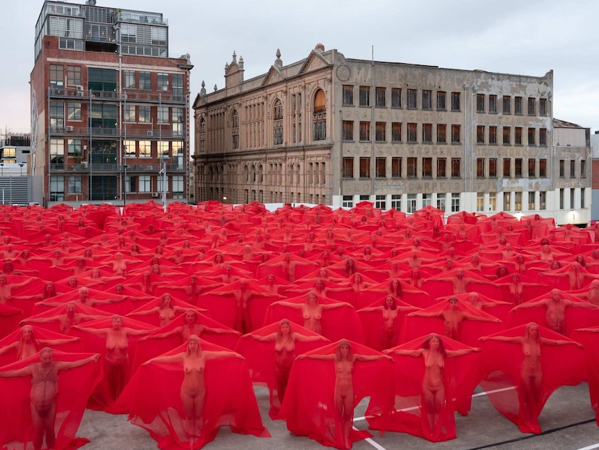 Naked people stand in a carpark under red veils.