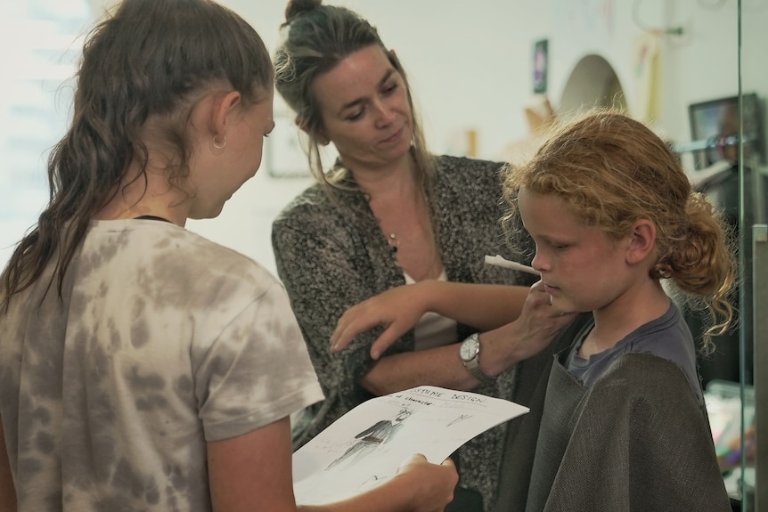 A teacher helps a young student fit a costume while another looks on holding the design.