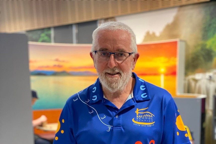 A man with short, white hair and goatie, wearing reading glasses is standing in front of a colourful mural, looking at camera.