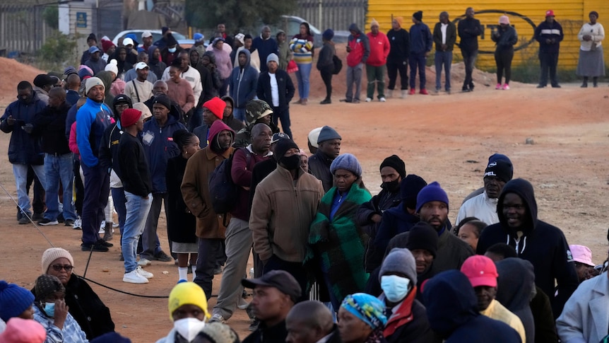 A long line of people stand in a snaking queue on red dirt