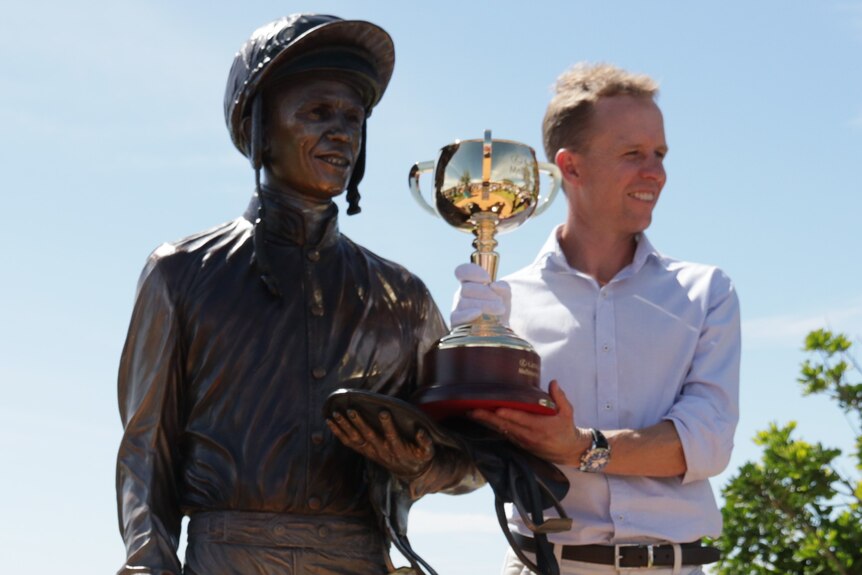Man wearing a white glove holding golden cup up and standing next to statue on left