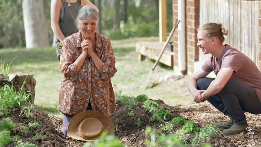 A lady kneeling down in a herb garden with a young man looking on, smiling.