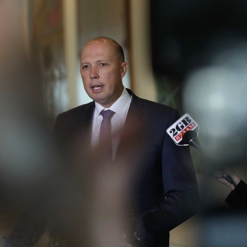 Peter Dutton is in focus in the background, in front of him are blurred reporters with microphones.