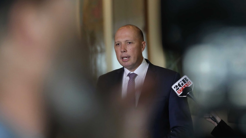 Peter Dutton is in focus in the background, in front of him are blurred reporters with microphones.