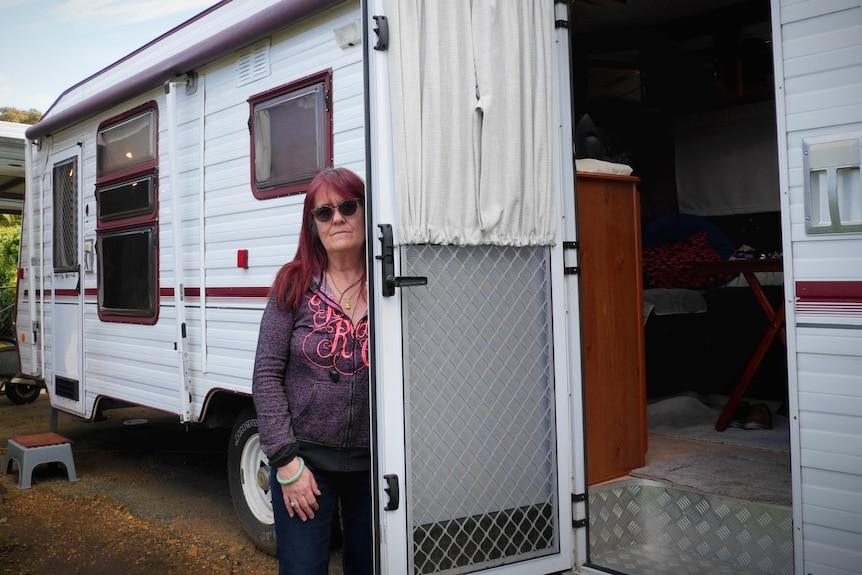 An older woman with red hair, wearing sunglasses, stands in front of a caravan.