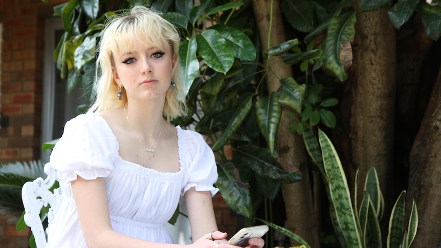 Dakota wearing a white dress, sitting at a table outside and holding her phone.