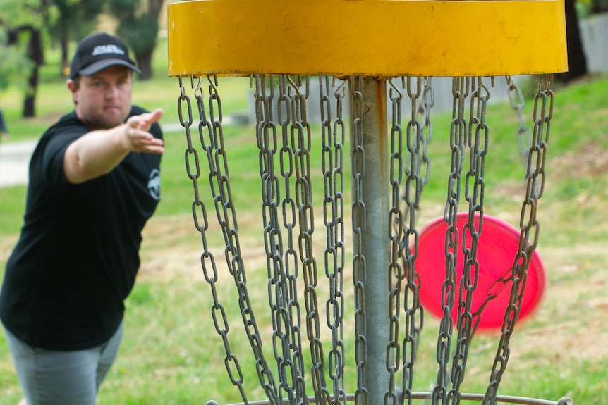 A man throwing a frisbee into a disc golf hole, a pole and basket with draping chains to catch the disc