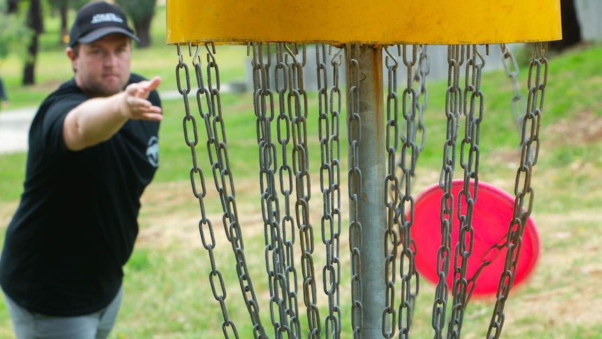 A man throwing a frisbee into a disc golf hole, a pole and basket with draping chains to catch the disc