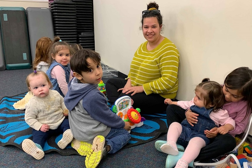 A small group of children are sitting on the floor playing with toys. A woman in a yellow and white top is sitting with them.