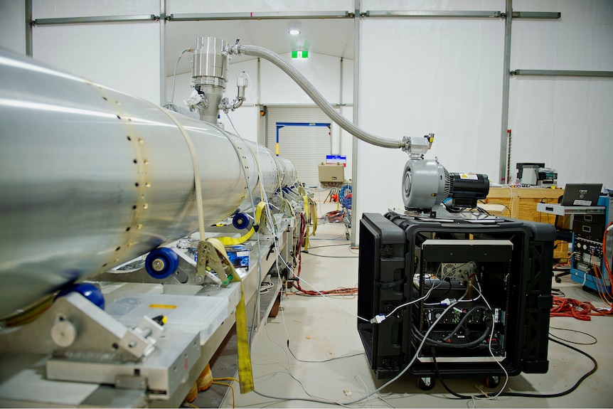 A long silver rocket is strapped to a table in a workroom with a large pump connected to it. 