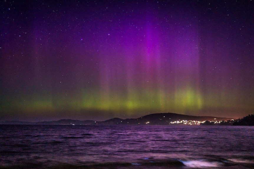Night sky lit up with pink and purple lights of the Aurora Australis and bioluminescence in the river.