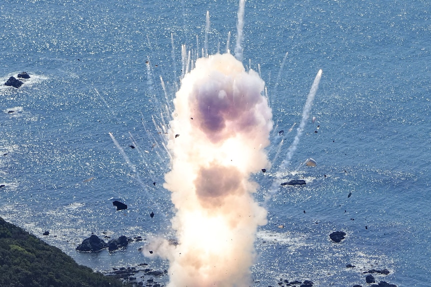 A vertically oriented explosion is visible against a backdrop of ocean and forested mountainside.