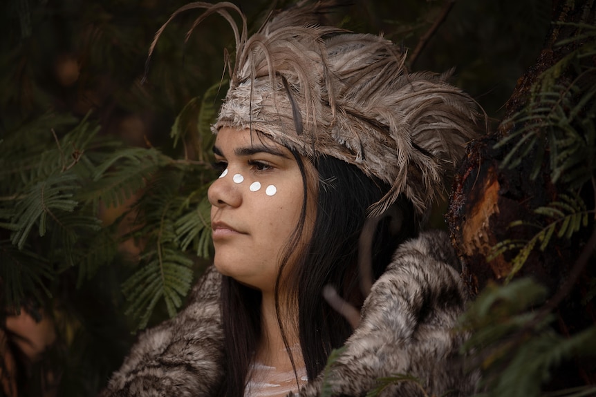 Profile of a young Aboriginal woman's face wearing a feathered head piece.