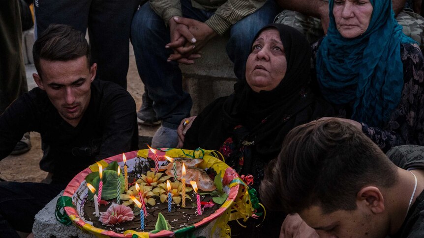 A woman in a black head scarf stare up at the sky, with candles and flowers in front of her. Others kneel on the ground.