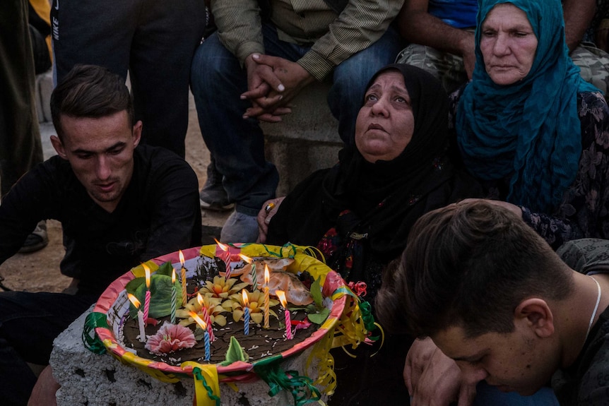 A woman in a black head scarf stare up at the sky, with candles and flowers in front of her. Others kneel on the ground.