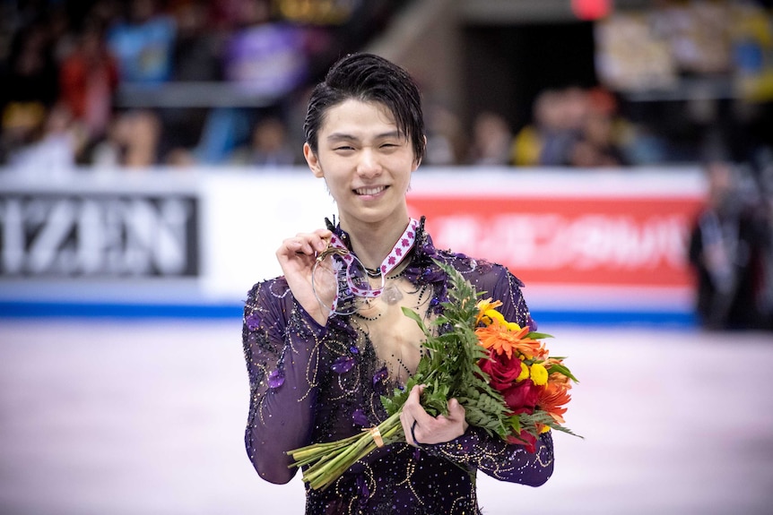 Yazuru Hanyu holds a medal and flowers and smiles towards the camera.