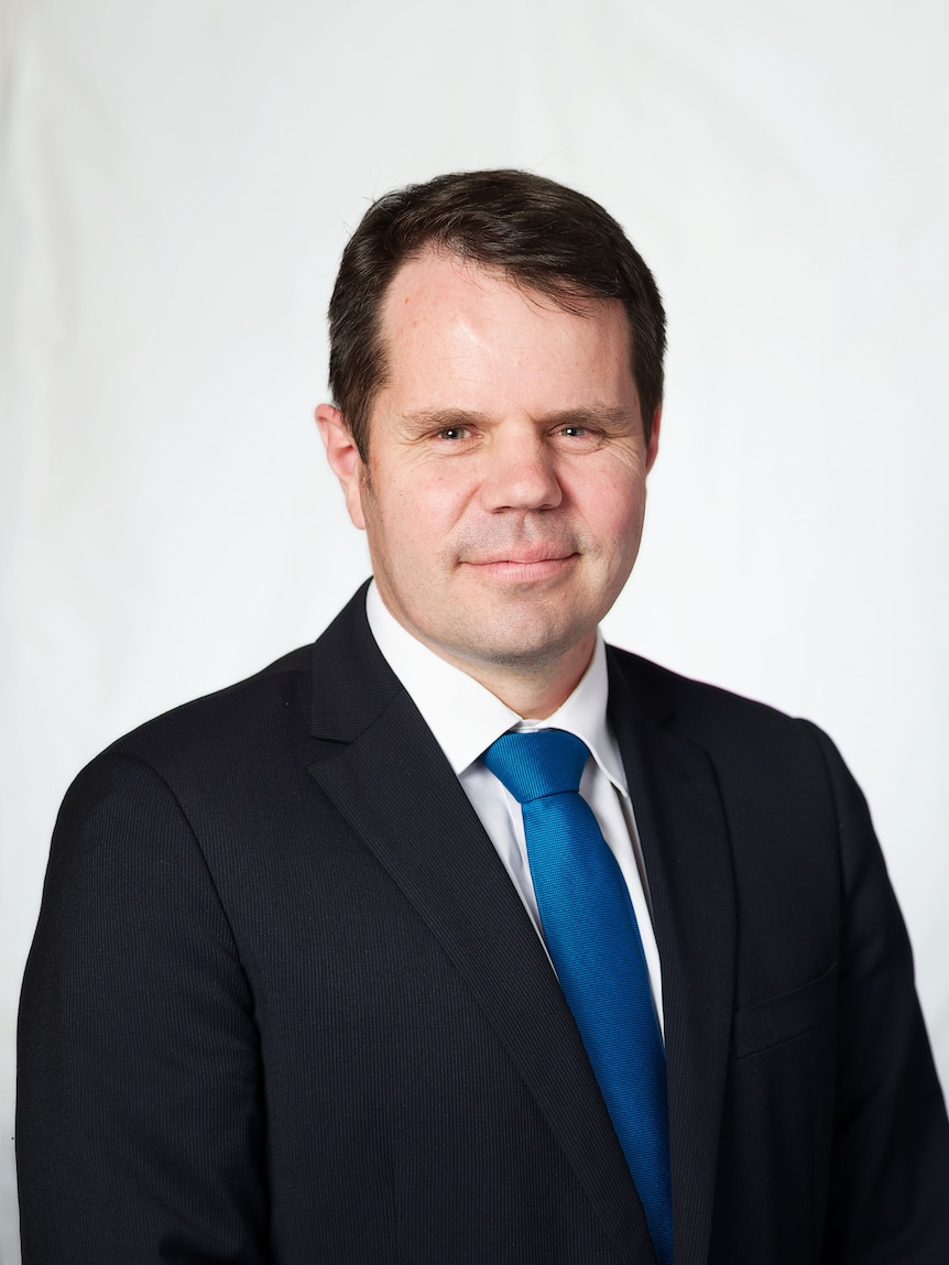 Man with brown hair wearing a black suit with a white shirt and blue tie.