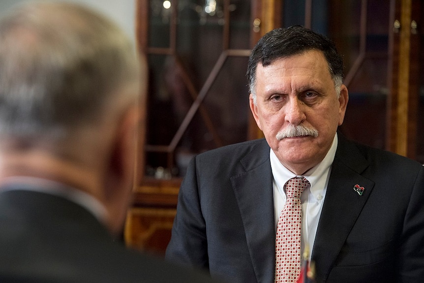 You look at Libya's Prime Minister Fayez Serraj in front of a brown cabinet from behind a man's shoulder who is blurred.