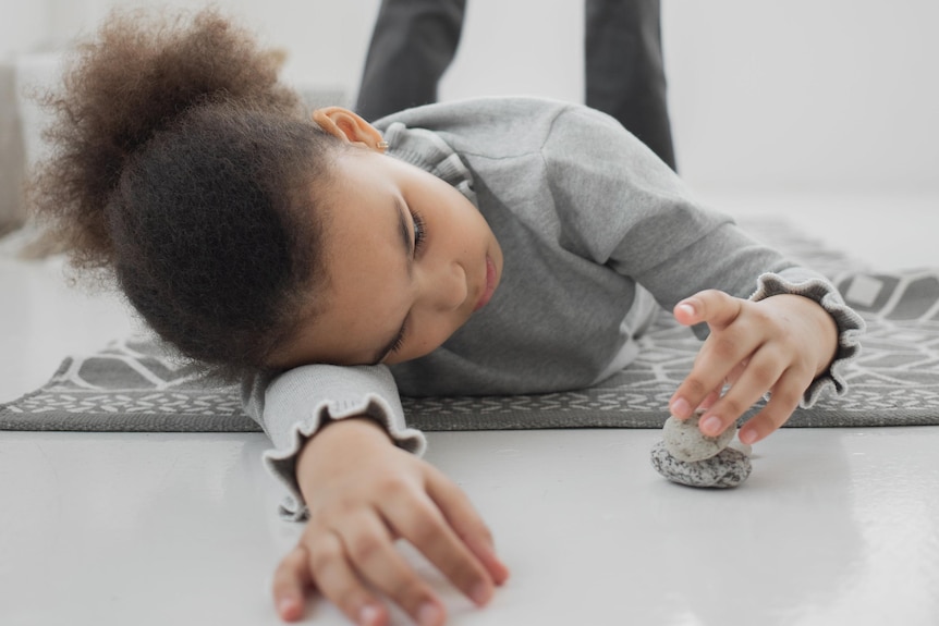 A child lies on the floor with a bored expression playing with stones. 