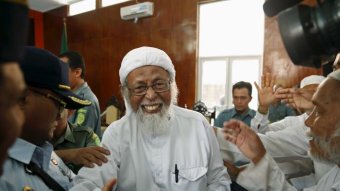 Abu Bakar Bashir smiles in a court room. He is wearing all white clothing.