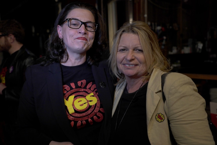 A woman wearing glasses and a t-shirt with Yes stands next to a blonde haired woman