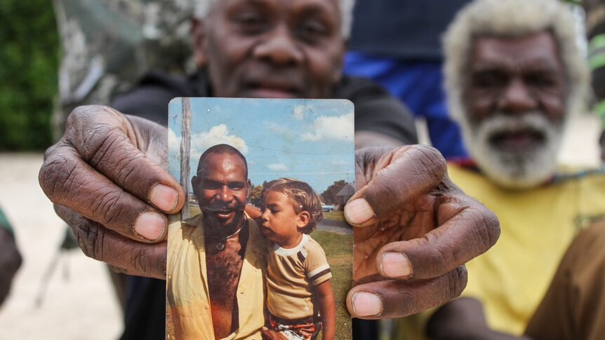 A man holds up a photo showing another man holding a toddler.