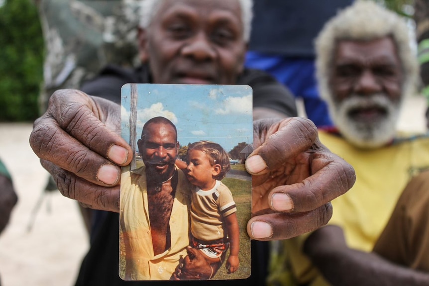 A man holds up a photo showing another man holding a toddler.