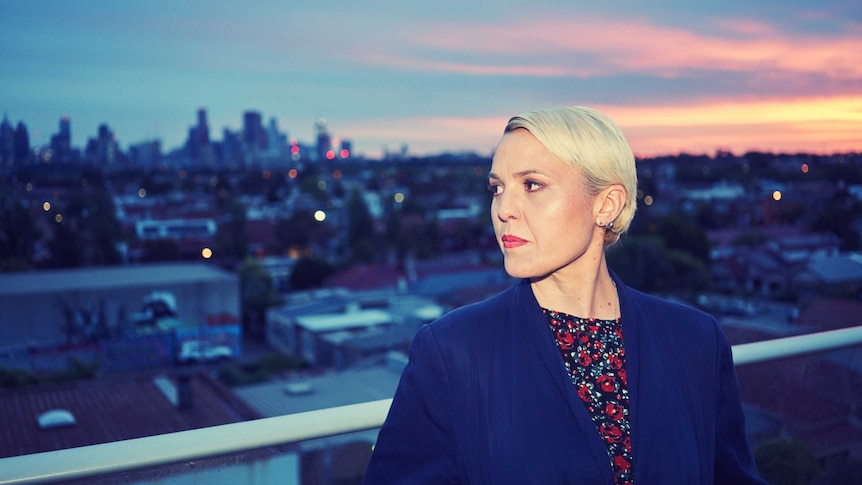 Liz stands on a balcony in front of a sunset. She's wearing a blue blazer and has short blond hair.
