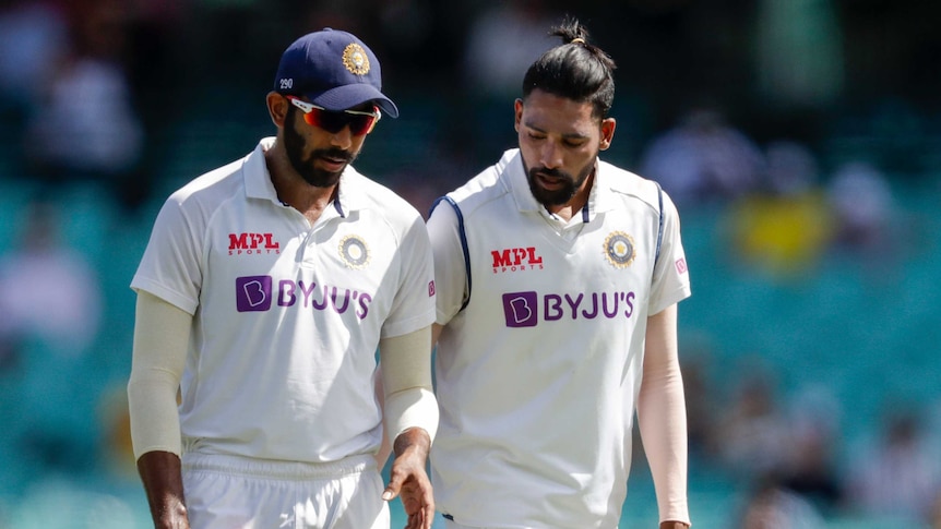 Two Indian players in cricket whites talk while walking on the field.