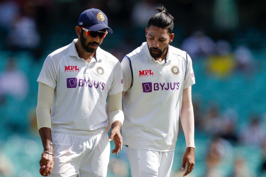 Two Indian players in cricket whites talk while walking on the field.