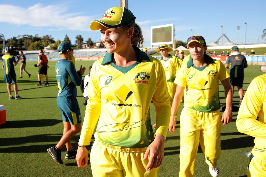 A woman wearing a cricket helmet, gloves and shirt stares intently into camera