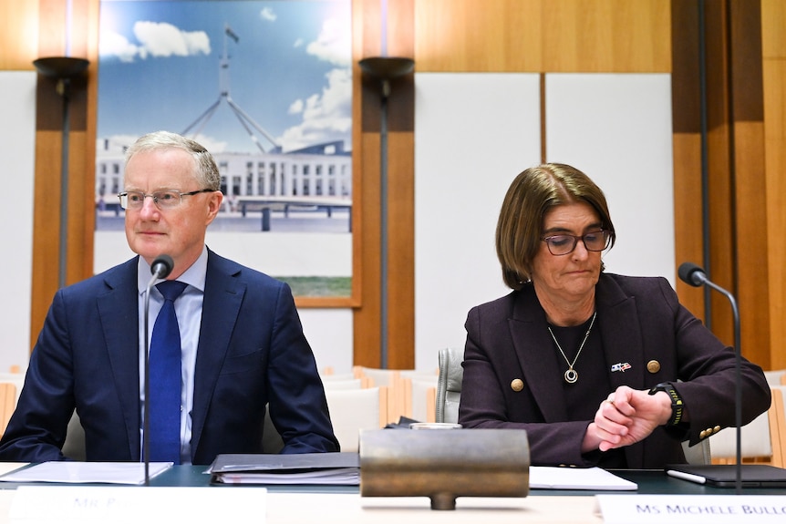 A man and woman sit at a table with small microphones in front of them. The woman is looking at a watch on her left hand.