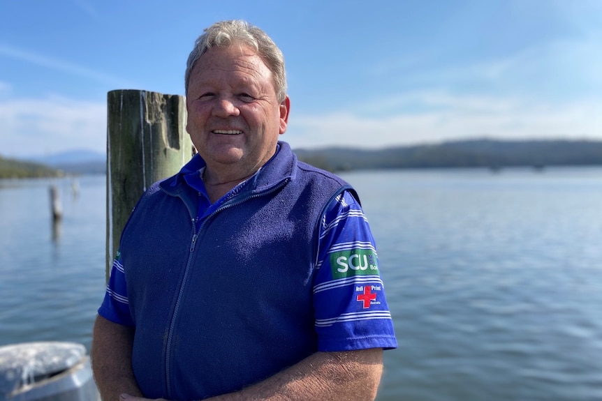 A man stands with a blue vest on a jetty with a river in the background.