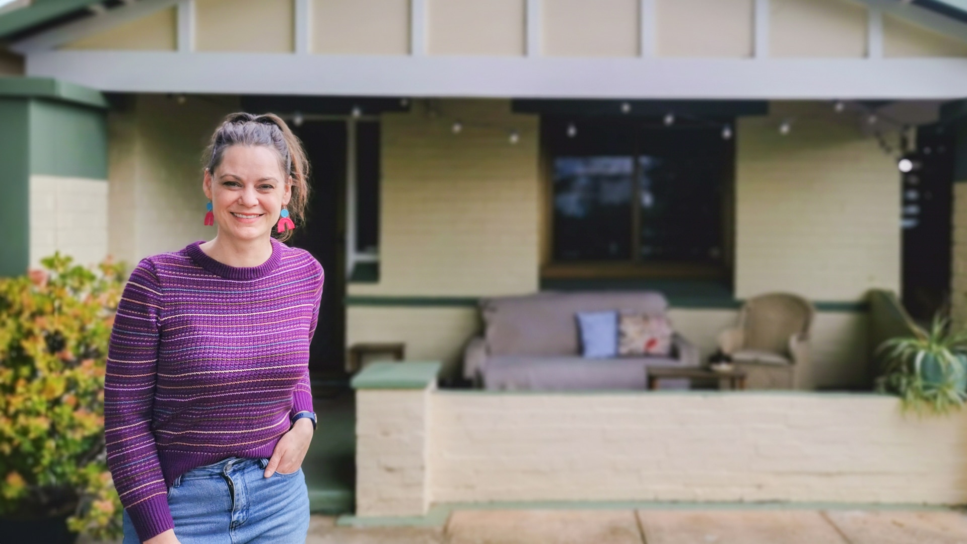 Koren smiles and stands in front of her house in Adelaide which has a cream and pale green facade.