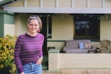 Koren smiles and stands in front of her house in Adelaide which has a cream and pale green facade.