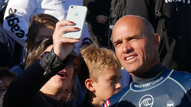 Kelly Slater takes a selfie photo in front of a group of fans.