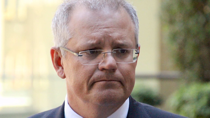 Prime Minister Scott Morrison looks to the left in portrait image. A reflection is visible in his glasses and he wears a suit.