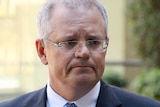 Prime Minister Scott Morrison looks to the left in portrait image. A reflection is visible in his glasses and he wears a suit.