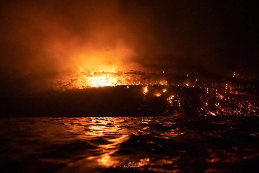 A fire burns on the headland, lighting up the trees, as it also spills down into the water.
