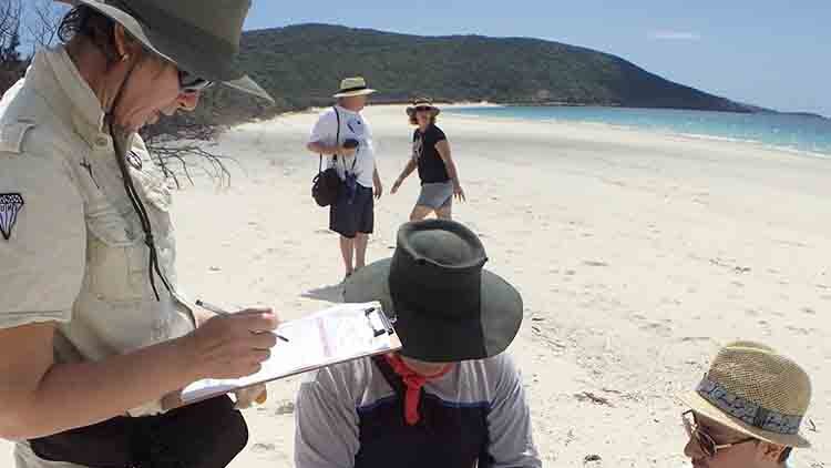 A woman stands on beach holding a clipboard while two men kneel placing rubbish in a garbage bag