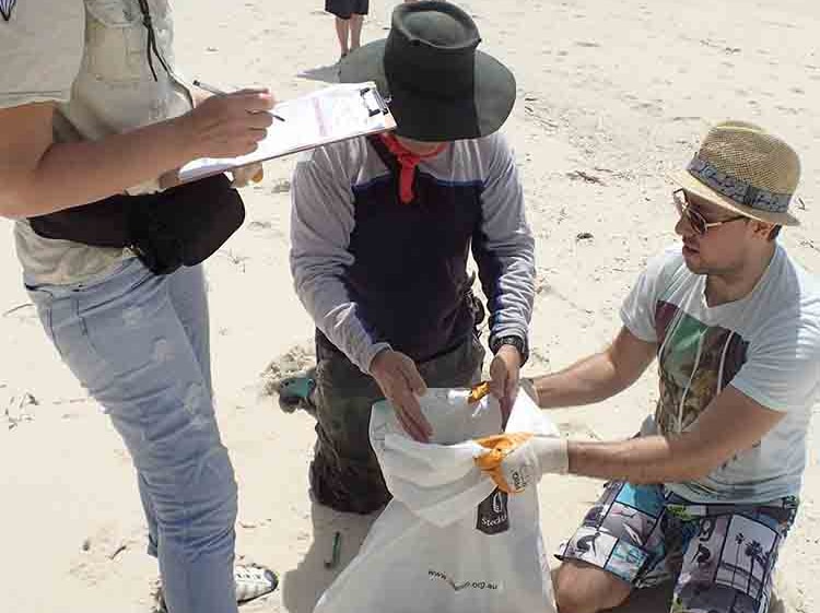 A woman stands on beach holding a clipboard while two men kneel placing rubbish in a garbage bag